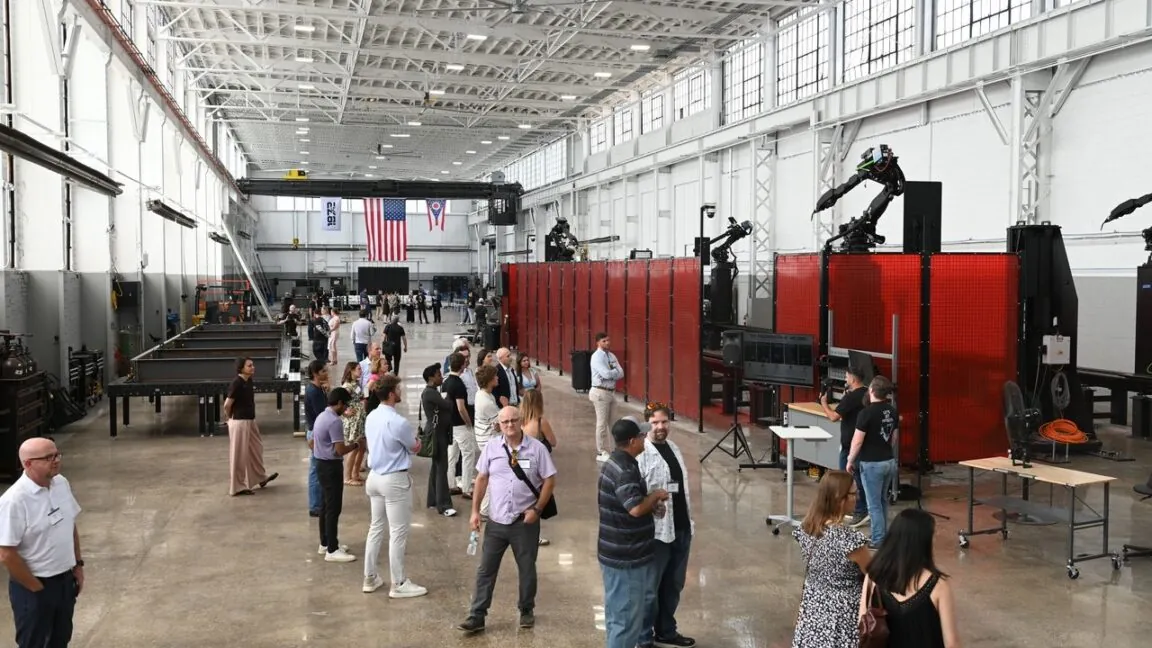 The inside view of a new factory opened by the startup 1872 on July 22, 2026. Welding workstations and robotic arms line the sides of the building while people mill around in the middle. A large American flag hangs against the back wall next to smaller flags representing the state of Ohio and the company 1872 logo.