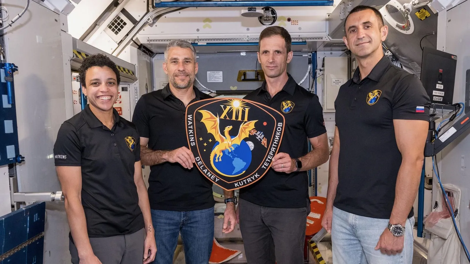 three men and one woman, all wearing black polo shirts, hold a plaque inside a mockup space module