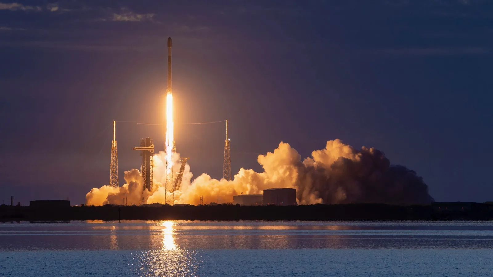 a black-and-white spacex falcon 9 rocket launches into a darkening evening sky.