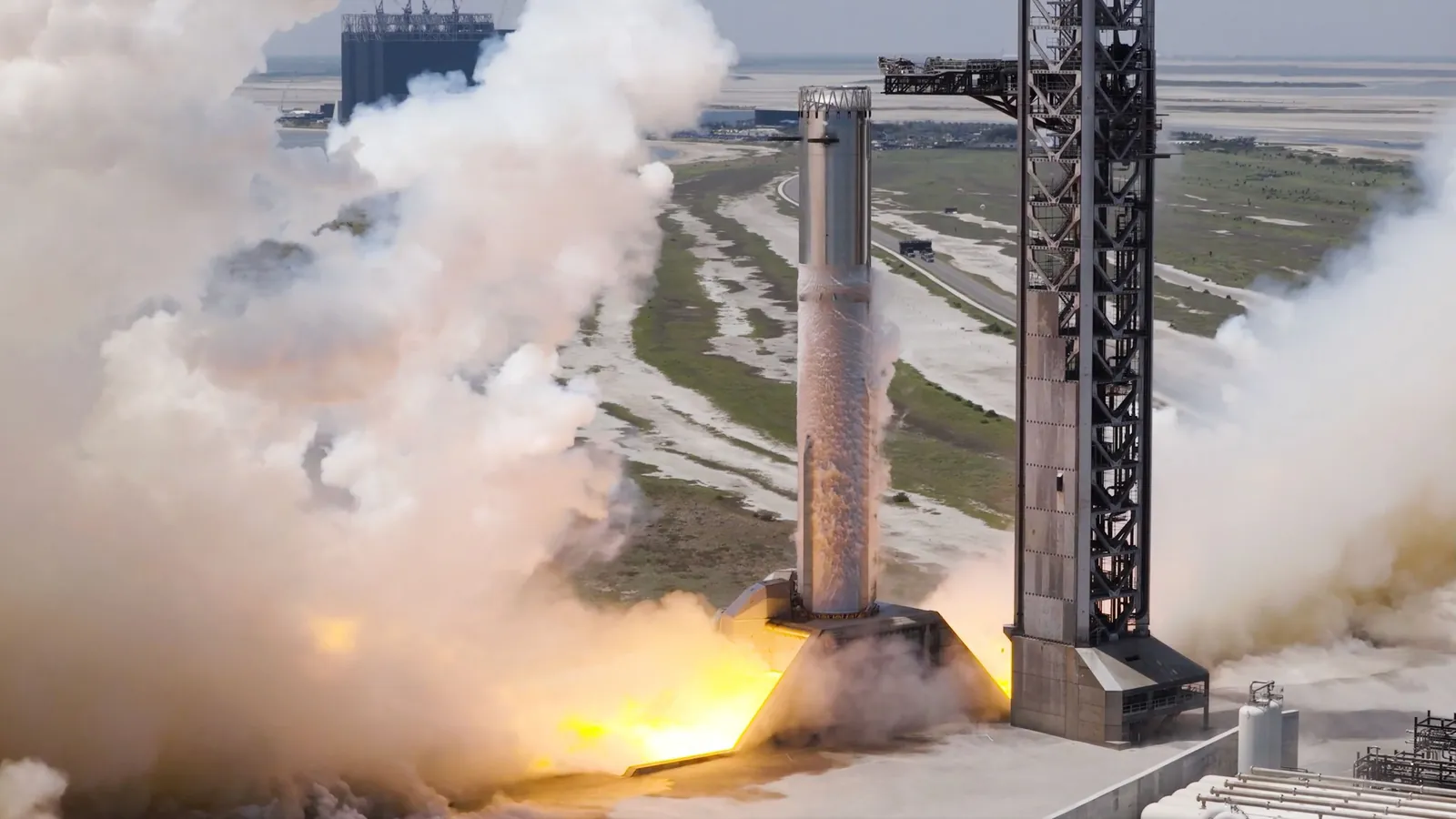 a large silver rocket conducts an engine test at a seaside launch pad